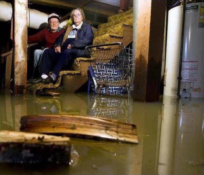 An older couple sitting on their basement stairs surrounded by standing water